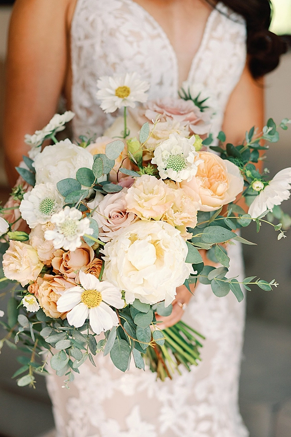 Bridal bouquet with peony wedding bouquet blooms, peach roses, daisies, and eucalyptus held against a lace wedding dress indoors