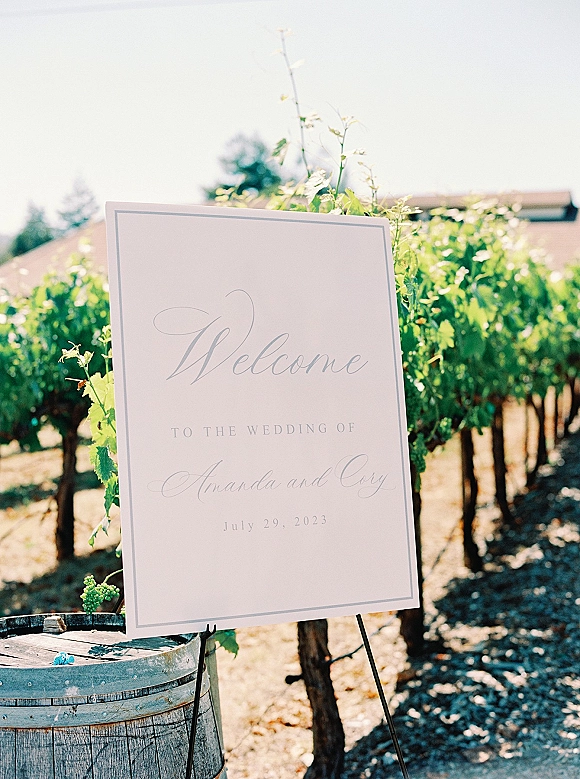 Wedding welcome sign with calligraphy lettering on a stand beside a wooden barrel, set along a gravel path with vineyard rows behind