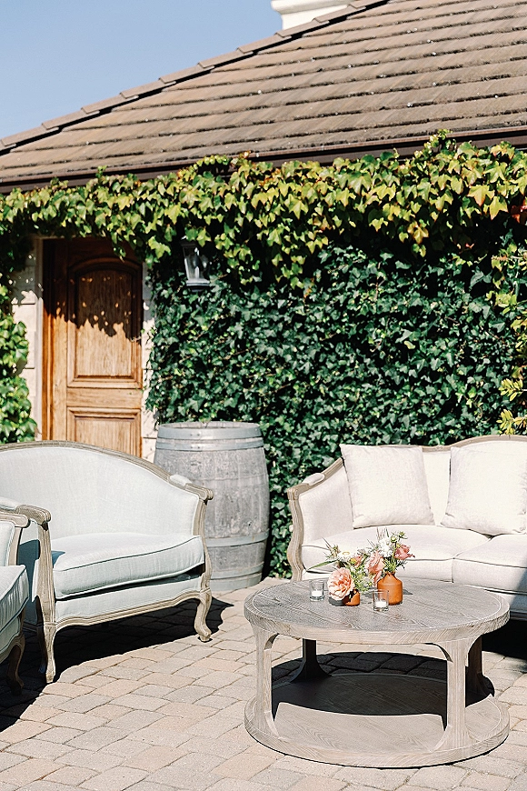 Wedding lounge seating with an outdoor wedding lounge sofa, upholstered chairs, and round coffee table with flowers, ivy wall behind