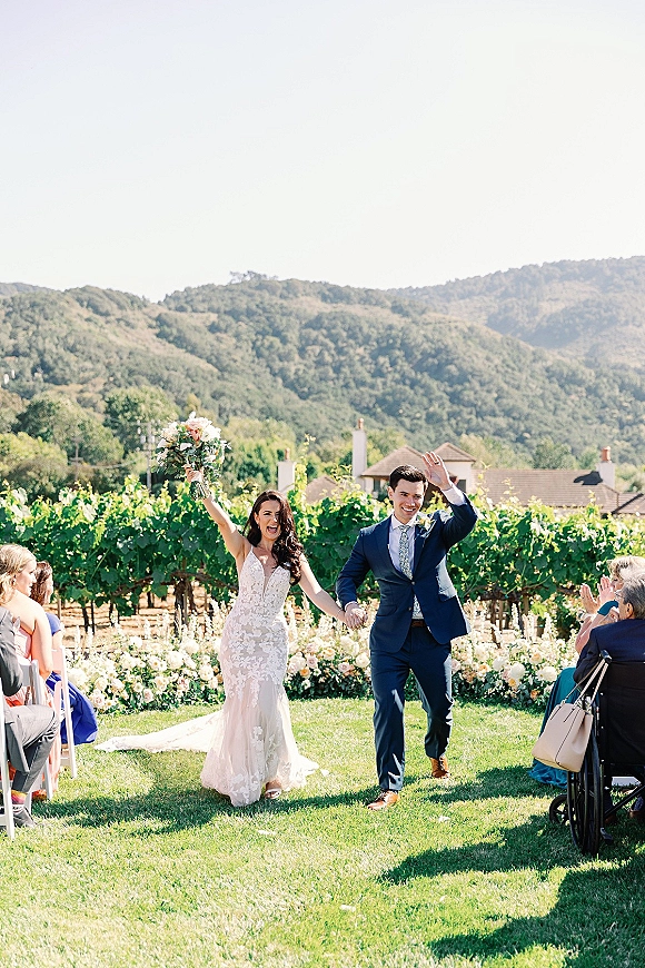 Wedding recessional as bride and groom hold hands, she lifts her bouquet overhead while guests watch along a floral-lined vineyard aisle.