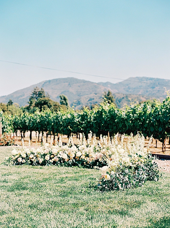 Ceremony floral decor with ground aisle flowers of roses, delphinium, and greenery lining a lawn aisle with vineyard rows and mountains beyond