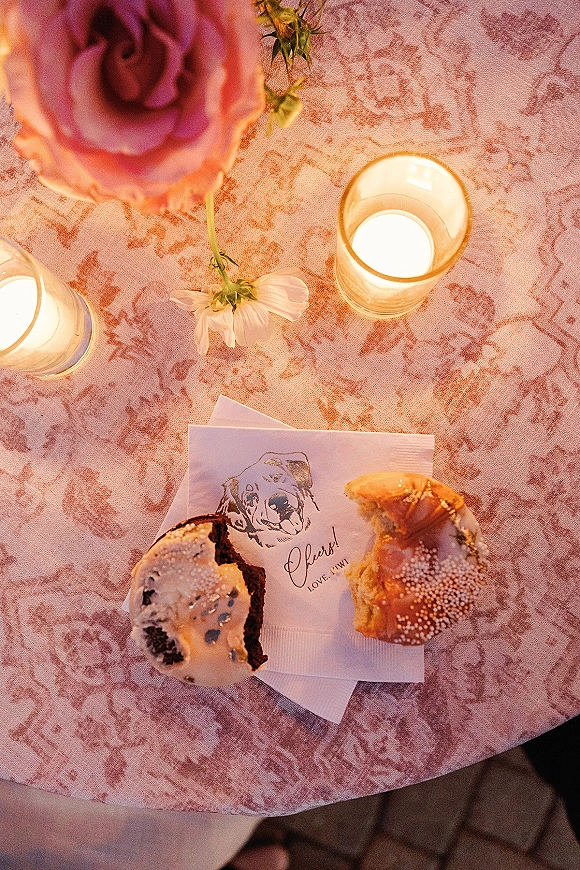 Wedding dessert table with glass votive candles, pastries dusted with powdered sugar, and a pink rose on a pink patterned tablecloth