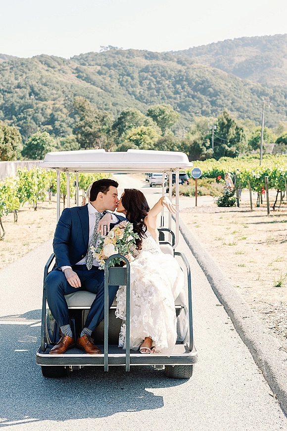 Wedding kiss portrait of bride and groom golf cart exit as they kiss on a vineyard road, bouquet in hand with mountains beyond