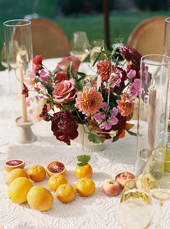 Wedding centerpiece with colorful floral centerpiece in a pedestal bowl, taper candles, citrus fruit, and glassware on lace table outdoors