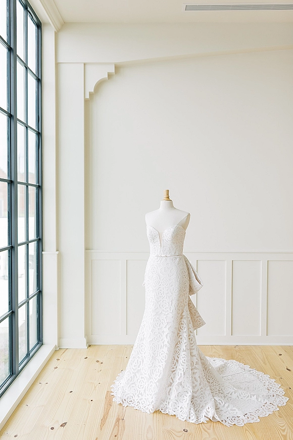 Wedding dress with strapless lace design on a mannequin, featuring a sweetheart neckline, side bow, and train in window light