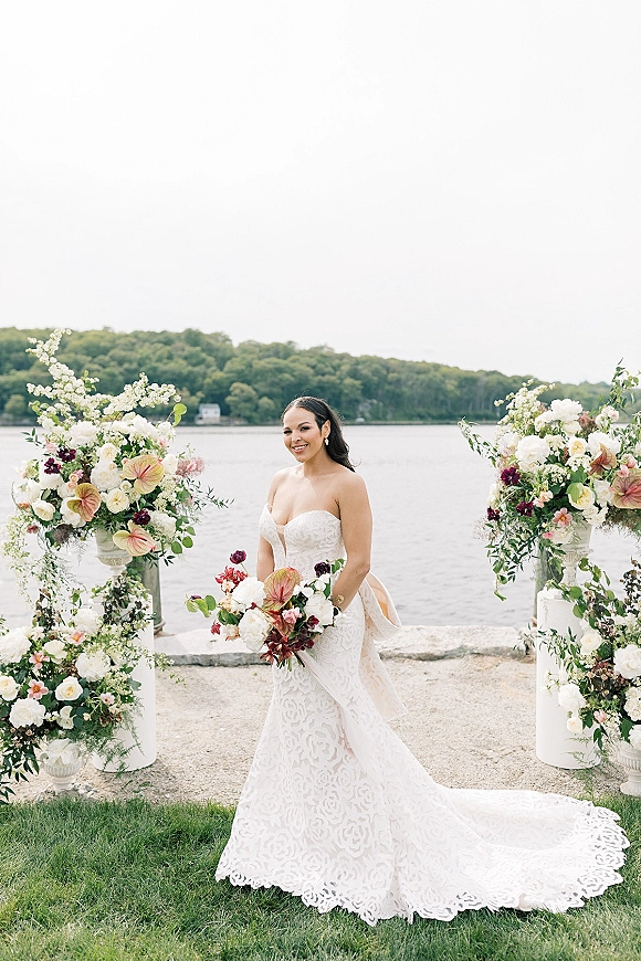 Bridal portrait of a bride in a strapless lace wedding dress with long train, holding a bouquet by a lakeshore with floral urns
