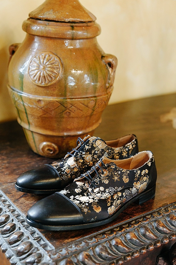 Groom shoes with floral groom shoes pattern, black leather toe caps and laces, styled on a carved wooden table beside a ceramic vase