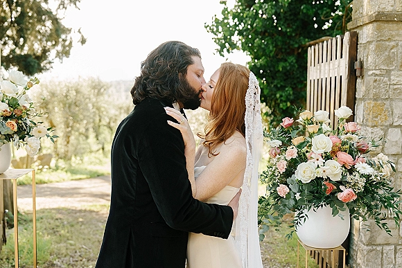 Wedding kiss as the bride and groom kiss beside rose and greenery urns, her lace veil and strapless dress against a stone wall backdrop