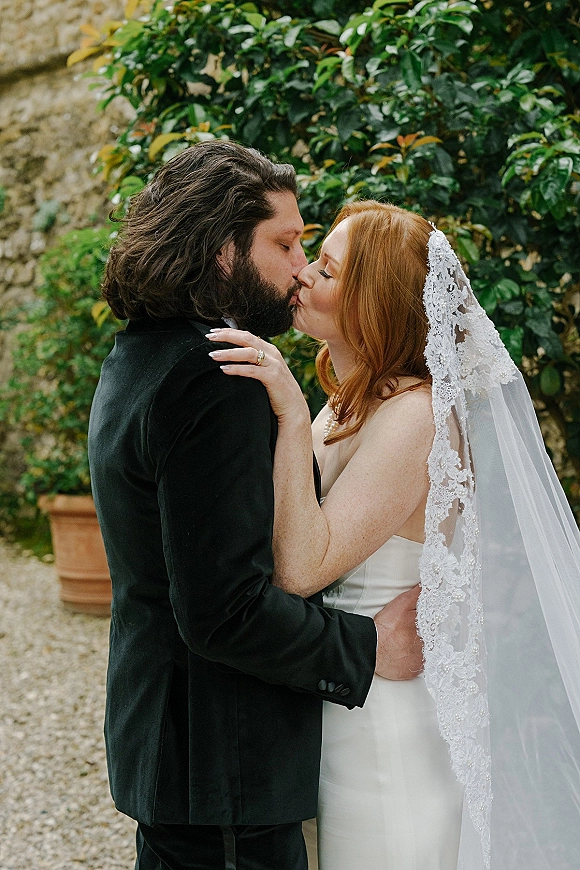 Wedding kiss portrait of bride and groom kissing close up, lace veil and strapless dress against green foliage and stone wall backdrop