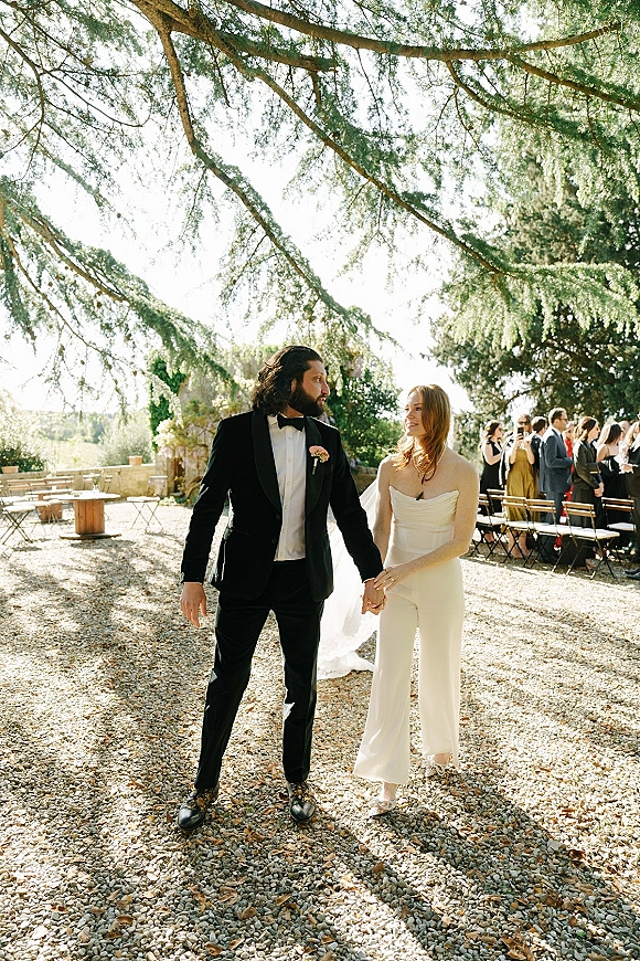Couple portrait of bride and groom holding hands, strapless dress and veil with black tuxedo under tree branches in a garden setting