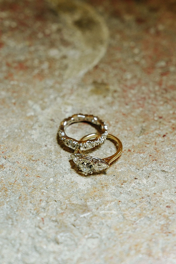Wedding rings with an engagement ring close up featuring a diamond accent, stacked with pavé and gold bands on a stone surface, glassware blurred behind