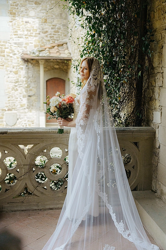Bridal portrait of a bride holding bouquet in a strapless wedding dress with long lace veil, looking over her shoulder in an ivy stone courtyard