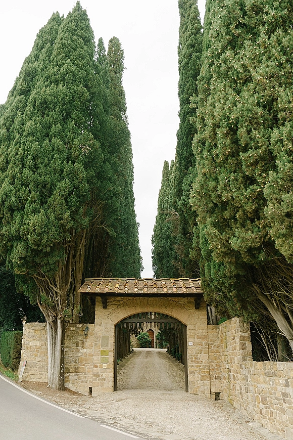 Wedding venue entrance with a stone gate entrance and wooden doors beneath a clay tile roof, framed by tall cypress trees along a gravel drive