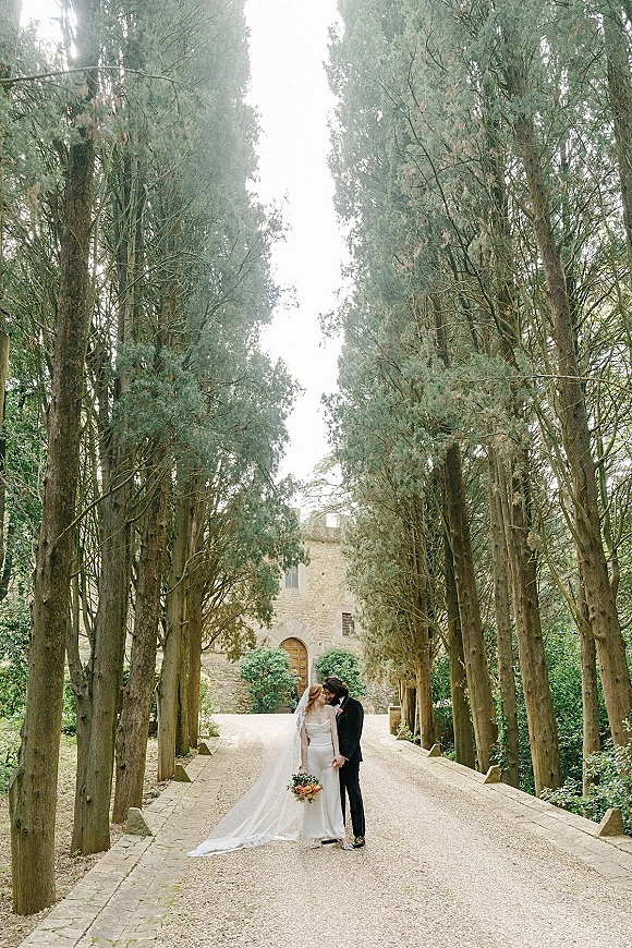 Couple portrait of bride and groom kiss while holding hands, her long veil and bouquet flowing down a tree-lined gravel drive by stone building.