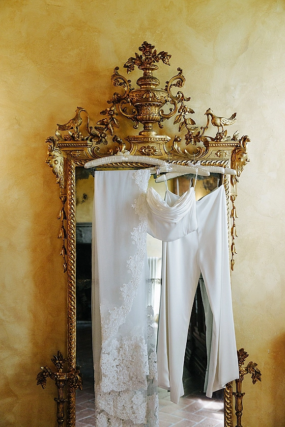 Bridal outfit with wedding getting ready details, lingerie set and lace veil on hangers beside an ornate gilt mirror on a plaster wall