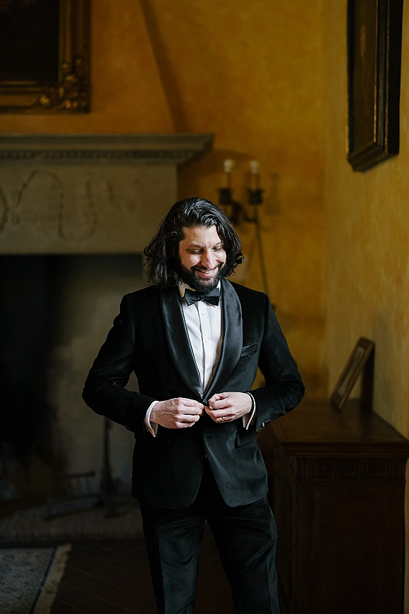 Groom portrait in a black tuxedo with bow tie and satin lapels, standing in an indoor room by a fireplace mantel and framed art