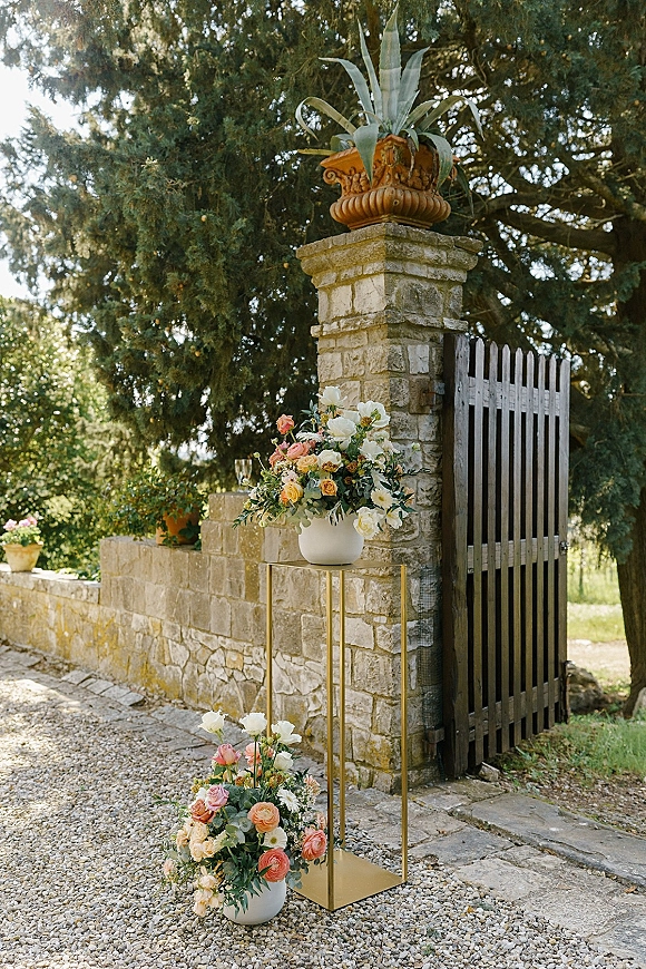 Wedding entrance decor with roses and greenery in white vases on gold pedestal stands, framing a wooden gate by a stone wall on gravel path