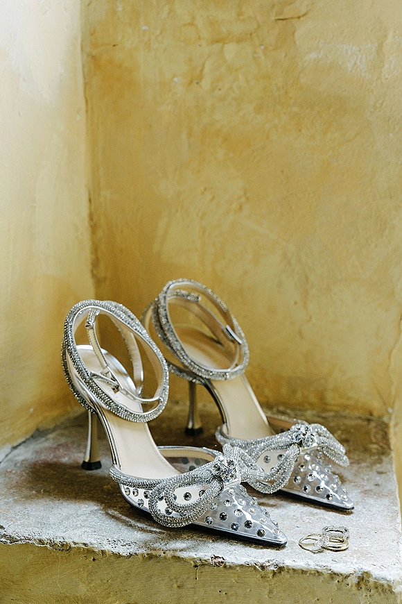 Bridal shoes with rhinestone bridal heels, crystal bow and clear ankle strap beside wedding rings on a stone ledge, plaster wall behind