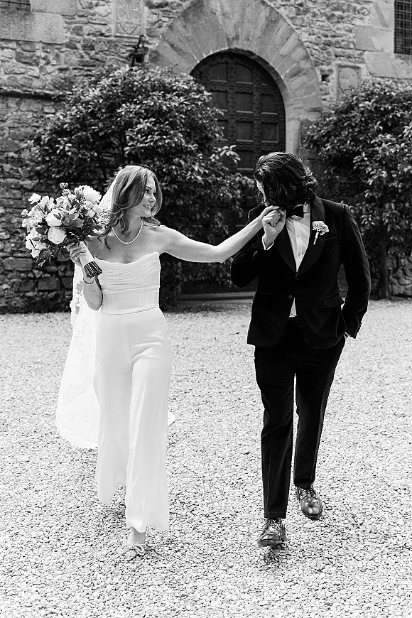 Couple portrait in a black and white wedding portrait style, groom kissing bride’s hand as she holds a bouquet by an arched stone doorway