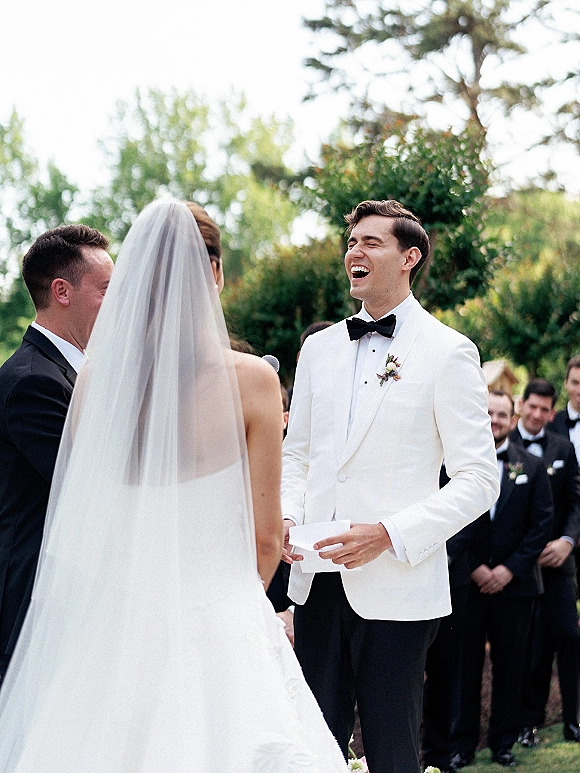 Wedding vows as groom reading vow cards laughs beside veiled bride in white dress, with guests and groomsmen amid trees behind
