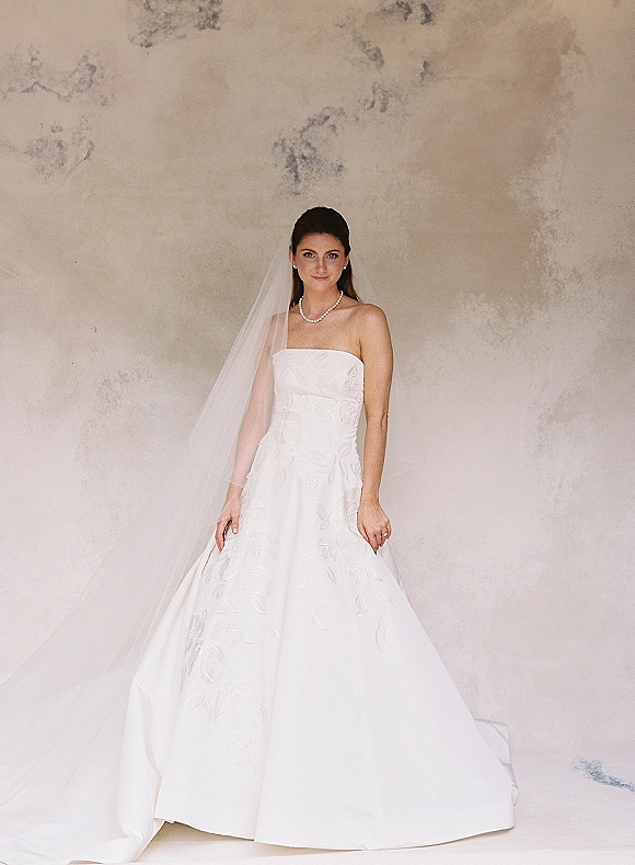 Bridal portrait of a bride in a strapless wedding dress with floral applique and cathedral veil, wearing a pearl necklace against a textured plaster wall