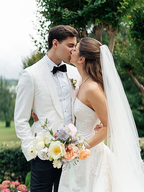 Wedding kiss portrait of bride and groom kissing outdoors, her long veil and bouquet visible, his white dinner jacket against garden greenery