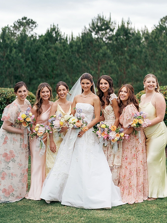 Bridal party portrait of bride with bridesmaids holding bouquets, bride in strapless gown and veil with pearl necklace on a lawn by trees