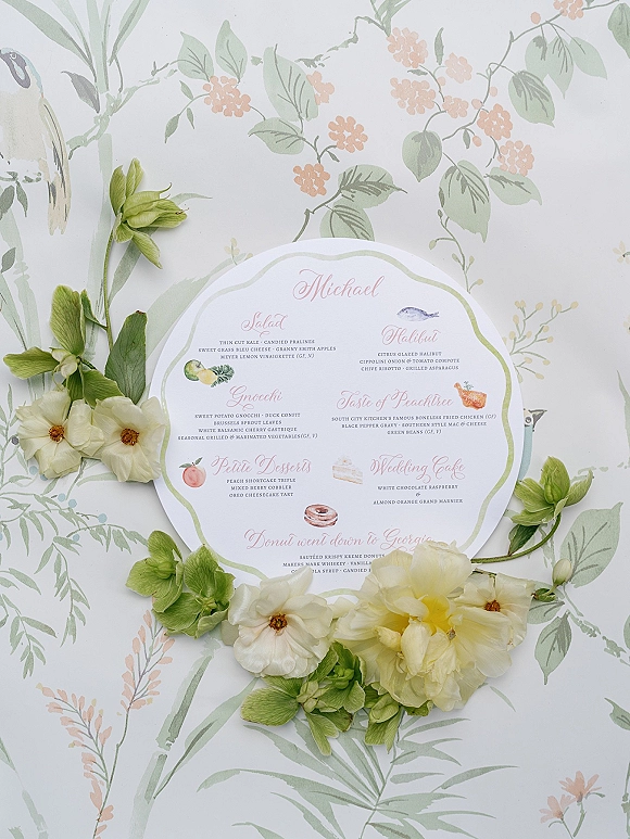 Wedding menu card with scalloped edges and floral illustration of white blooms and green foliage on light floral patterned paper flat lay