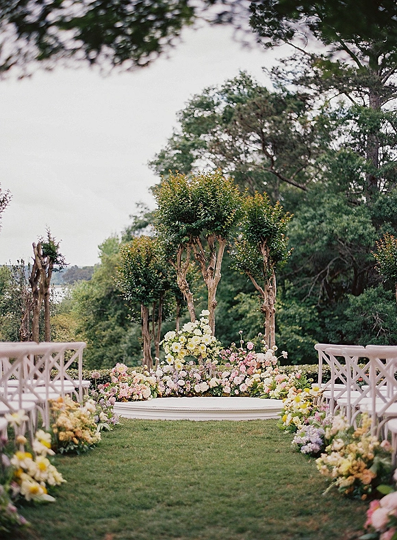 Outdoor ceremony setup with floral lined ceremony aisle, white crossback chairs, and a raised platform on a garden lawn by distant water