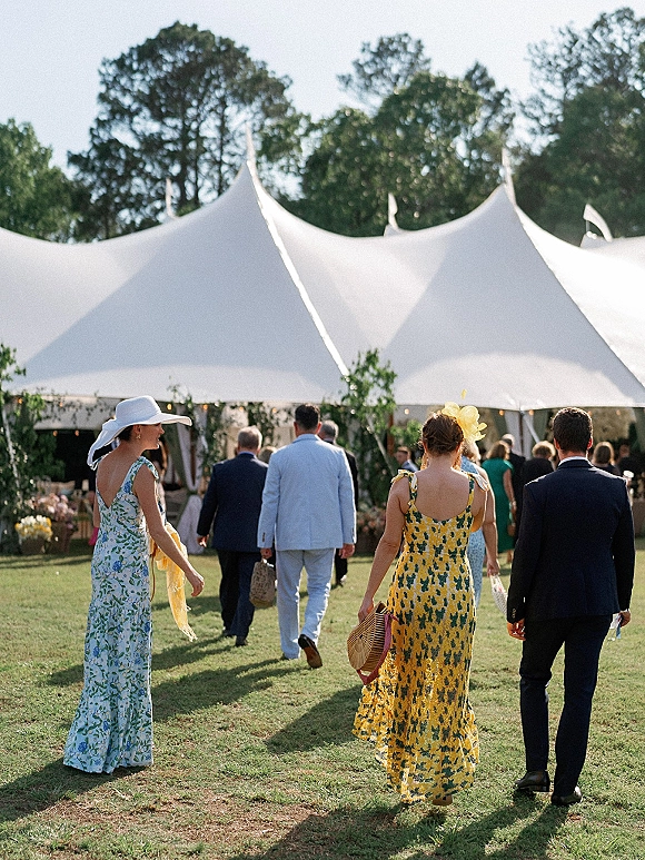 Wedding guests in floral dresses and navy suit stroll across a lawn, with a white sunhat and fascinator under a string-lit tent