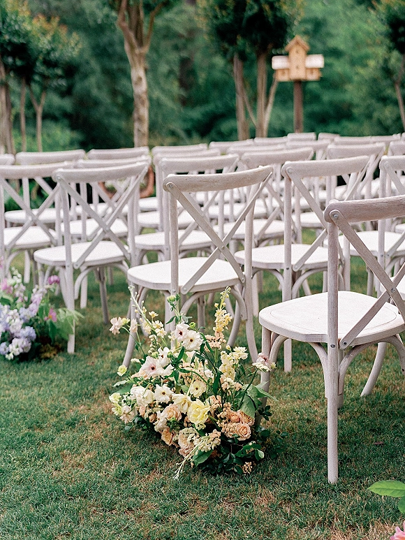 Ceremony seating with outdoor ceremony chairs in neat rows of white crossback chairs, lined with aisle flowers on a garden lawn under trees