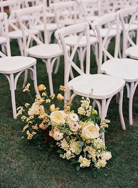 Ceremony aisle decor with white cross back chairs and low cream rose and yellow floral ground arrangement on a grassy outdoor lawn
