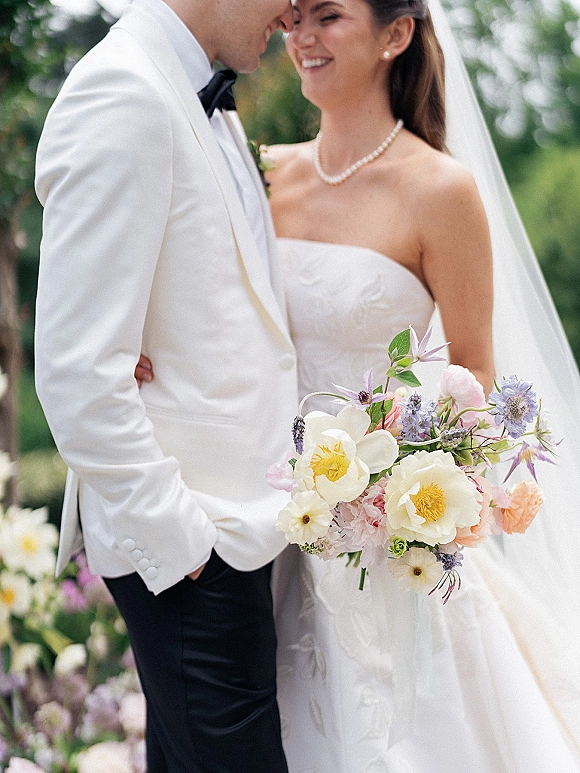 Couple portrait of bride in strapless wedding dress and veil with pearl necklace, groom in white tuxedo, holding pastel bouquet in garden greenery