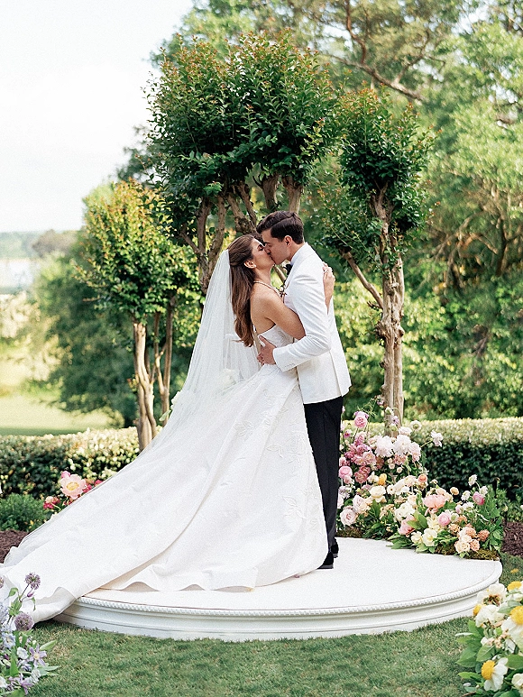 Wedding kiss portrait of bride and groom kissing on a round white platform, veil and long train in a garden with hedges and water view