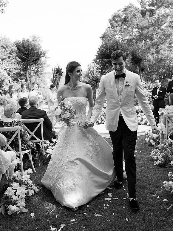 Wedding recessional as bride and groom walk the aisle hand in hand, petals underfoot, bouquet and veil flowing in an outdoor garden setting
