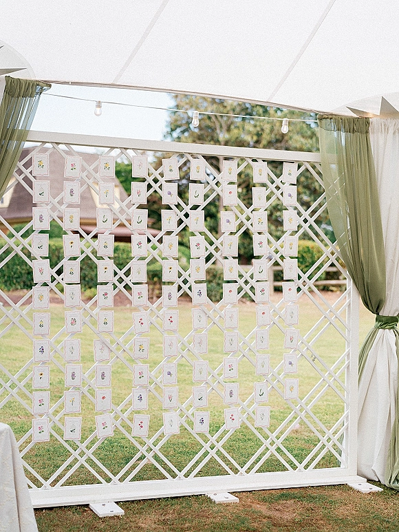 Seating chart display with wedding escort card display on a white lattice backdrop, draped sage curtains and string lights in a garden lawn setting