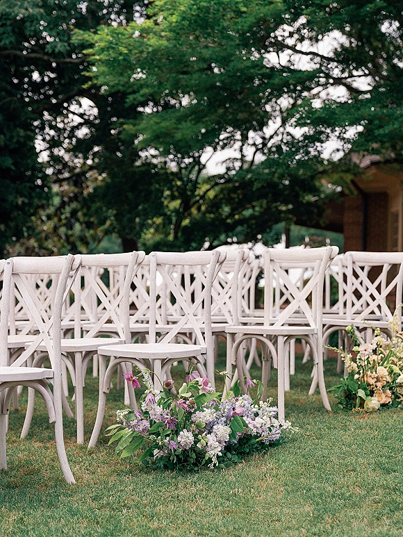 Ceremony seating with outdoor wedding chairs in neat rows, lined by purple-and-white floral aisle clusters on a grassy lawn under trees