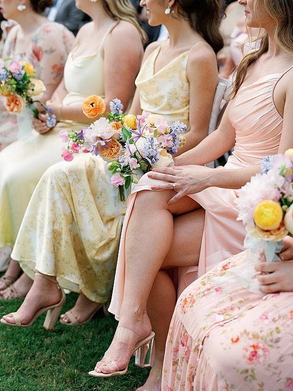 Bridesmaids seated holding bouquets in pastel bridesmaid dresses, statement earrings and heeled sandals, with guests behind on lawn chairs outdoors