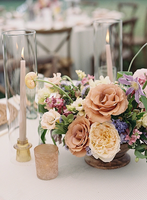 Wedding centerpiece with a low floral centerpiece of roses, mixed blooms, and greenery, surrounded by taper candles in glass hurricanes on a linen tablecloth
