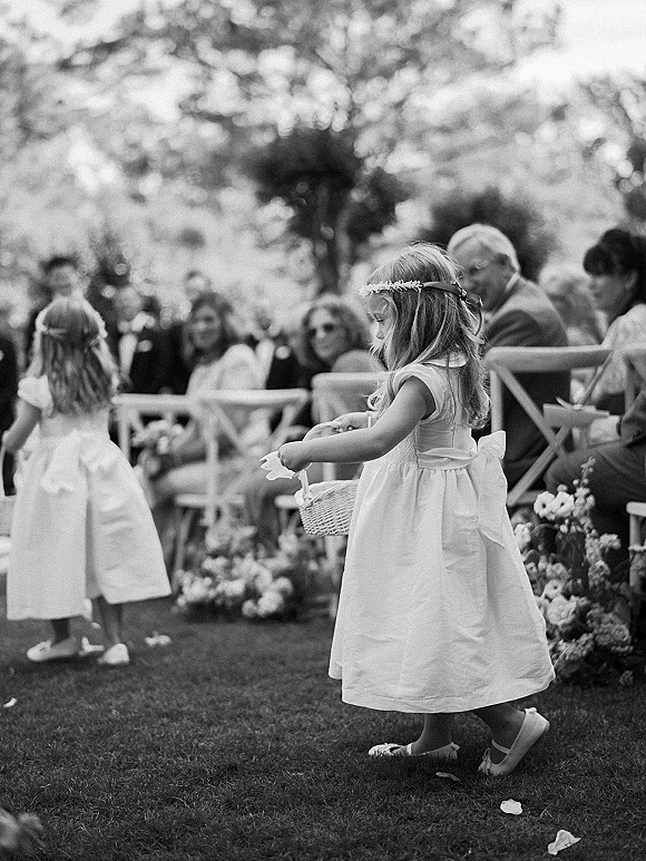 Flower girl walking aisle with basket, wearing a white dress and flower crown, petals underfoot on an outdoor lawn by guests
