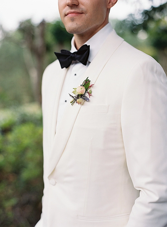 Groom portrait in a white tuxedo jacket with a black bow tie and pink ranunculus boutonniere, standing by lush outdoor greenery.