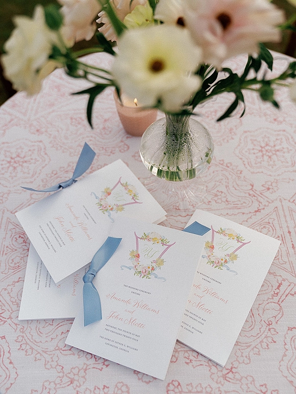 Wedding invitation suite with floral wedding invitations, blue ribbon, and calligraphy beside a bud vase on a pink patterned tablecloth