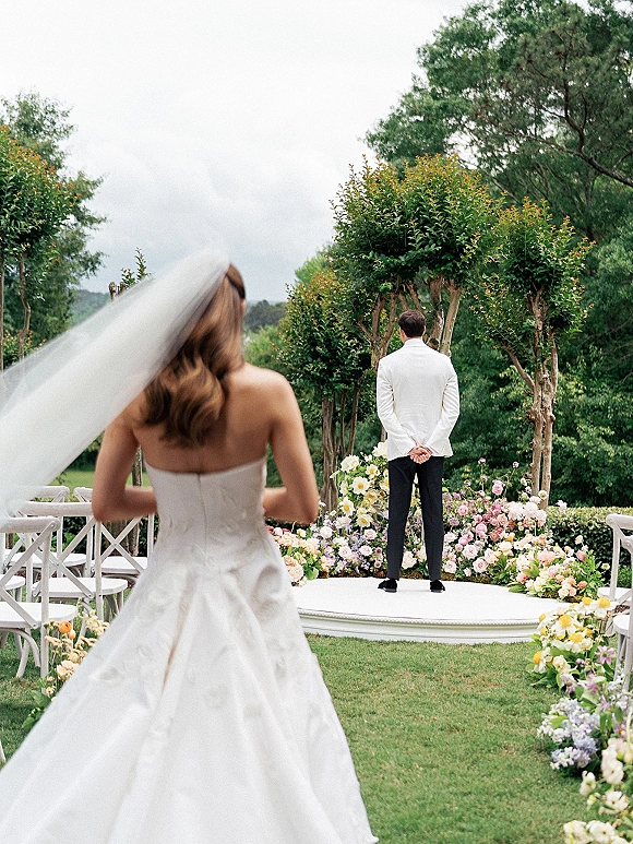 Wedding first look as bride walks to groom in an outdoor first look, veil trailing down a floral-lined garden aisle under cloudy skies