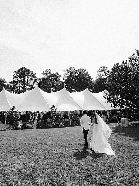 Newlywed couple walking hand in hand, bride’s cathedral veil trailing, toward a sailcloth tent reception with flags on a lawn