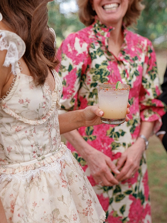 Wedding cocktail in a guest’s hand with a salted chili rim and lime wedge, held by a pink floral lace-trim dress at outdoor lawn hour