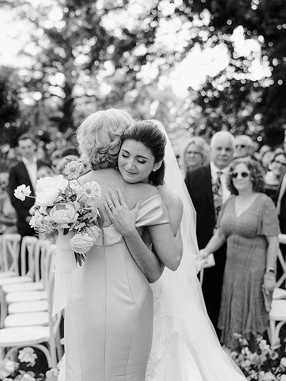 Wedding hug as bride embraces her mother, bouquet and veil visible, with ceremony chairs, guests, and trees in the background