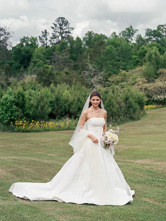 Bridal portrait of a bride in a strapless ball gown holding a bouquet, veil flowing, standing in a grassy field with wildflowers under clouds