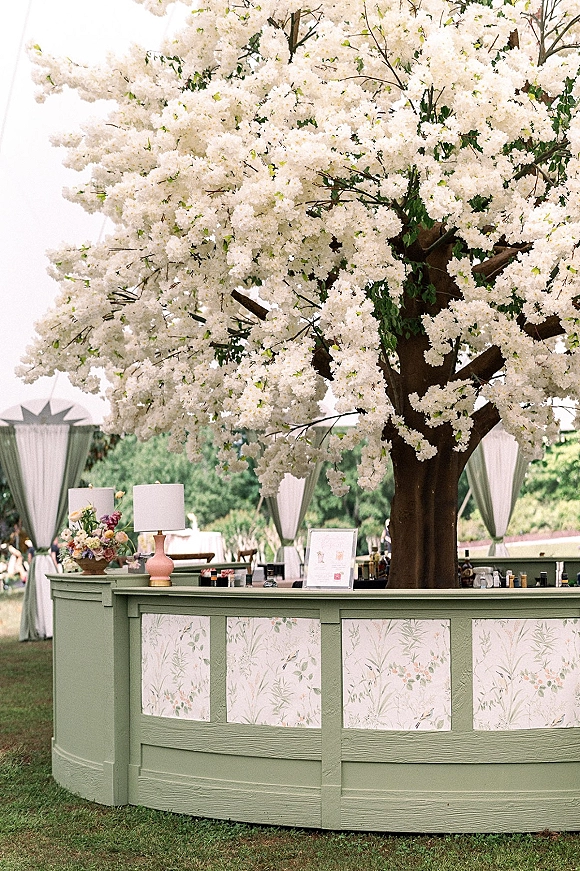 Wedding bar setup with a blossom tree installation over a curved bar counter, bottles and glassware, plus draped cabanas on a lawn
