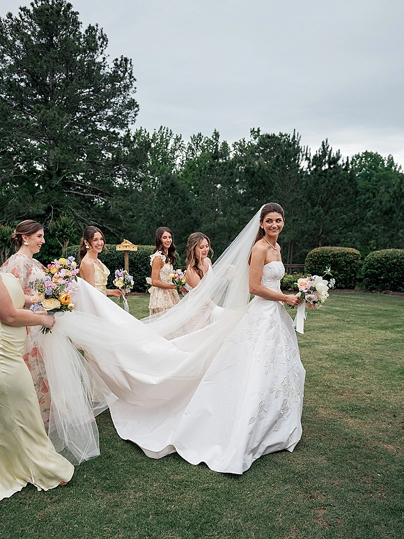 Bride with bridesmaids walking as they lift her cathedral veil and train, bouquets in hand, across a green lawn with trees under cloudy sky