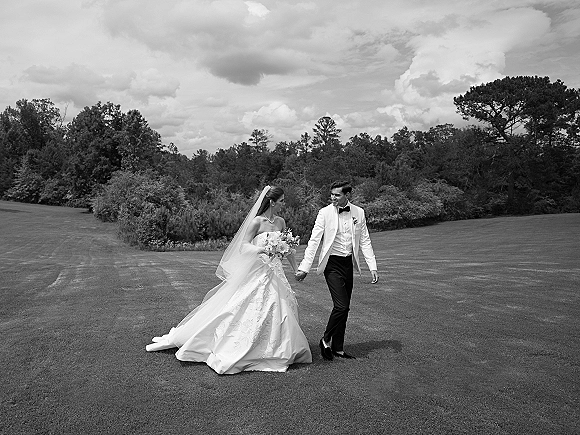 Couple portrait of bride and groom holding hands, walking on a grassy lawn under cloudy sky, bride with veil and bouquet, groom in tuxedo
